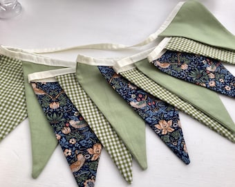May include: A fabric bunting banner featuring alternating triangular flags in solid sage green, white and green gingham check, and a dark blue floral pattern with birds and orange flowers, all attached to a continuous cream-colored fabric tape.