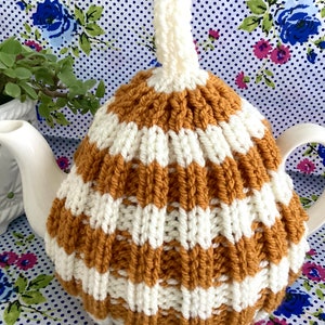 May include: A white teapot with a brown and white striped knitted tea cosy. The cosy has a loop on top for hanging.
