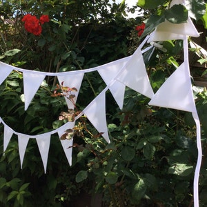 May include: A white bunting with triangular flags strung between green bushes. The bunting is made of fabric and has a white string.