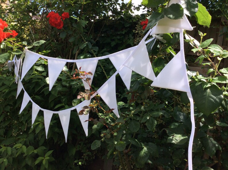 May include: A white bunting with triangular flags strung between green bushes. The bunting is made of fabric and has a white string.