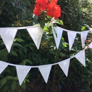 May include: A white bunting with triangular flags strung together. The bunting is hanging in a garden setting with green foliage and red flowers.