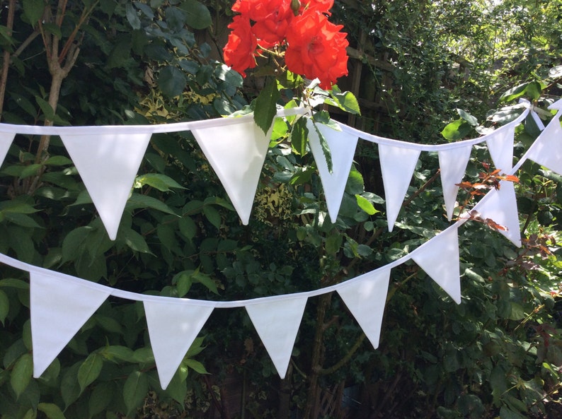 May include: A white bunting with triangular flags strung together. The bunting is hanging in a garden setting with green foliage and red flowers.