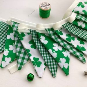 May include: A green and white fabric banner with shamrock designs. The banner is made of two different fabrics, one with a green and white gingham pattern and the other with white shamrocks on a green background. The banner is strung together with white ribbon.