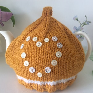 May include: A mustard yellow knitted teapot cozy with a white trim and white buttons arranged in a heart shape.