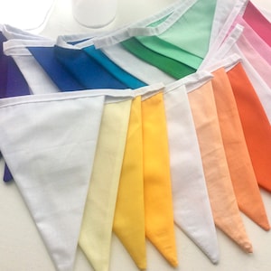 Peut inclure: Une guirlande de tissu arc-en-ciel avec des drapeaux triangulaires dans des tons de violet, bleu, vert, jaune, orange et rose. La guirlande est en coton et a une bordure blanche.