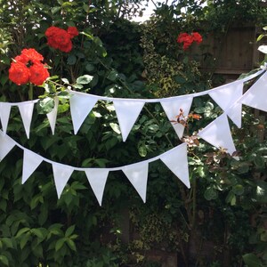 May include: White bunting with triangular flags strung between green bushes. The bunting is made of fabric and has a white string.