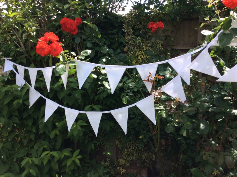 May include: White bunting with triangular flags strung between green bushes. The bunting is made of fabric and has a white string.
