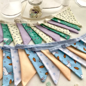 May include: Several colorful fabric bunting banners. The banners feature pastel colors and patterns, including bunnies, carrots, and stars. The bunting is made of triangles sewn onto a white fabric strip. A jar of buttons is in the background.