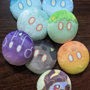 Puede incluir: Ocho juguetes de peluche redondos con diferentes diseños. Los juguetes tienen aproximadamente 7,5 cm de diámetro y presentan varios colores y patrones. Los diseños incluyen un remolino azul y blanco, un remolino verde y blanco, un remolino morado y blanco, un remolino gris y blanco, un remolino azul claro y blanco, un remolino verde claro y blanco, un remolino amarillo y naranja, y un remolino morado y azul con un personaje de dibujos animados.