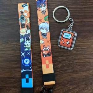 Op de afbeelding: Twee oranje en zwarte lanyards met cartoonfiguren en een zilveren sleutelhanger met een transparante plastic hanger. De hanger is een gamecontroller met een vinkje en een plusteken.