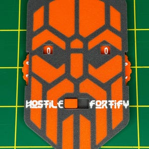 Può includere: Un pezzo di gioco grigio e arancione con le parole "HOSTILE" e "FORTIFY" stampate sopra. Il pezzo ha due quadranti che mostrano il numero "0". Il design presenta un motivo geometrico a forma di volto.