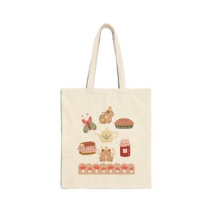 May include: A cream-colored canvas tote bag featuring a printed design of two frogs, a butterfly, a teapot, a pie, a jar of jam, and a loaf of bread. A horizontal border at the bottom displays a row of small red and white mushrooms on a tan background.