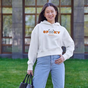 May include: A white cropped hoodie featuring the orange text "SPOOKY" with two small black eyes above the letters and the word "Season" printed in smaller orange font underneath, paired with light blue denim jeans and a black tote bag.