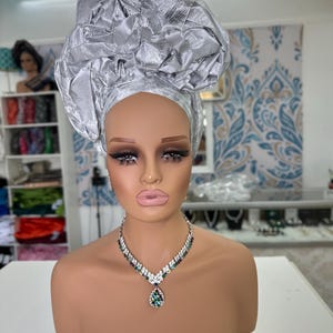 Può includere: Un busto di manichino con un turbante argentato e una collana scintillante con un pendente a goccia. La collana presenta pietre trasparenti e verde smeraldo. Il turbante è realizzato in un tessuto lucido e strutturato.