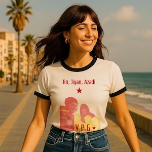 May include: Cream-colored ringer t-shirt with black trim featuring the text "Jin, Jiyan, Azadi" and a red star. The shirt has a graphic of people and the letters "YPG". The person is wearing blue jeans.