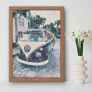 May include: A vintage blue and white Volkswagen van with a surfboard on the roof, parked in front of a white building with a green door. The van has American flags hanging from the side mirrors. The image is painted in a watercolour style.