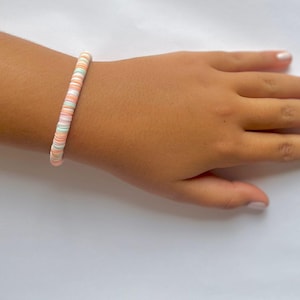 Pode incluir: Um bracelete de contas coloridas com contas rosa, brancas e verdes. O bracelete é feito de pequenas contas redondas que são enfiadas em um cordão elástico fino.