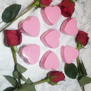 May include: Eight pink heart-shaped soaps arranged on a white marble surface with red roses and green leaves.