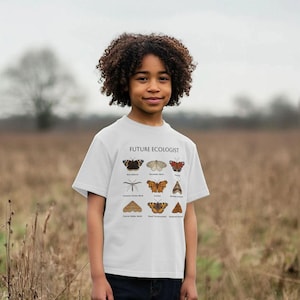 Peut inclure: Un t-shirt blanc avec l'inscription "FUTURE ECOLOGIST" et des illustrations de papillons et d'insectes. Le t-shirt est porté par une jeune personne aux cheveux bouclés, debout dans un champ.