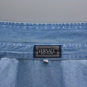 以下が含まれることがあります： 明るいブルーデニムのクローズアップ。ブランド名「VERSACE JEANS COUTURE」の白い文字が入った、暗い長方形のラベルが付いています。デニムにはステッチが見えます。
