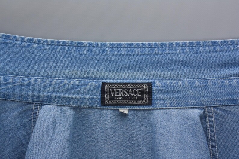 以下が含まれることがあります： 明るいブルーデニムのクローズアップ。ブランド名「VERSACE JEANS COUTURE」の白い文字が入った、暗い長方形のラベルが付いています。デニムにはステッチが見えます。