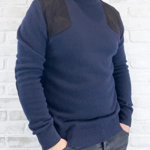 May include: Navy blue sweater with black shoulder patches. The sweater has a crew neck and ribbed cuffs and hem. The person is wearing dark gray jeans with their hands in their pockets. The sweater is a classic style.