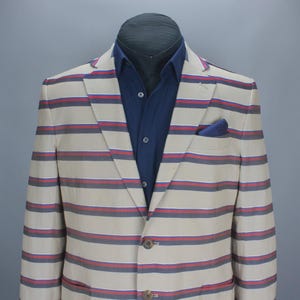 Peut inclure: Un blazer beige à rayures bleues marine et rouges, porté avec une chemise bleu marine et une pochette bleu marine à pois blancs. Le blazer est présenté sur un mannequin.