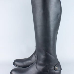 Può includere: Un paio di stivali da equitazione alti in pelle nera. Gli stivali hanno una finitura liscia, una punta arrotondata e un piccolo tacco. Una cerniera corre lungo la parte posteriore di ogni stivale. Accenti metallici decorativi sono visibili vicino alla caviglia e nella parte superiore dello stivale.