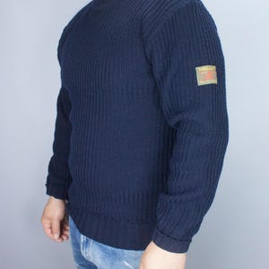 May include: Navy blue ribbed knit sweater with a crew neck and long sleeves. A small rectangular patch with text and a logo is on the upper left sleeve. The sweater is worn with blue jeans.