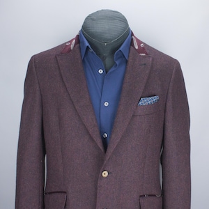 May include: A dark purple blazer with a textured finish, featuring a blue button-down shirt underneath. The blazer has a maroon paisley collar detail, two light-colored buttons, and a small blue patterned pocket square in the chest pocket.