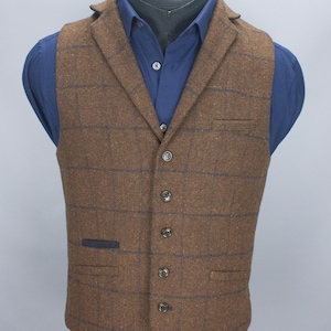 Puede incluir: Chaleco de tweed marrón con un patrón de cuadros azules, usado sobre una camisa abotonada azul. El chaleco tiene solapas con muescas, cierre de cinco botones y dos bolsillos delanteros. La prenda se exhibe en un maniquí.