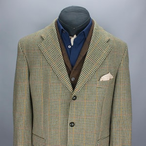 May include: A tweed blazer with a brown vest and a blue button-down shirt. The blazer has a classic houndstooth pattern in shades of green, brown, and yellow. A patterned pocket square is visible in the chest pocket.