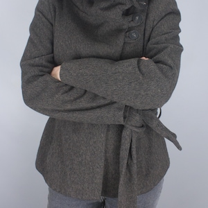May include: A dark gray hooded coat with a wrap-around collar and a tie belt. The coat has a textured appearance and is buttoned on the side. The sleeves are long, and the person is wearing gray pants.