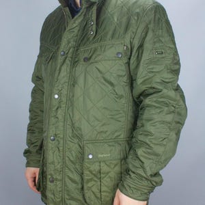 May include: Olive green quilted jacket with a diamond pattern. Features a stand-up collar, multiple pockets, and a button and zipper closure. The jacket has the brand name "Barbour" on a pocket.