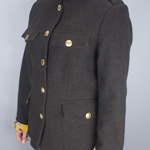 Puede incluir: Chaqueta de estilo militar gris oscuro con botones dorados y detalles de bolsillo. La chaqueta tiene un cuello alto y detalles decorativos en los hombros. Los puños están forrados con un estampado floral colorido.