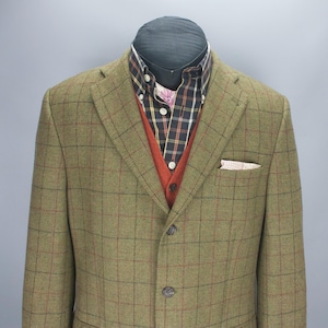 Puede incluir: Una chaqueta de tweed verde con un patrón de cuadros rojos y negros, combinada con un chaleco color óxido y una camisa a cuadros. Un pañuelo de bolsillo es visible en el bolsillo del pecho de la chaqueta. El conjunto se muestra en un maniquí.