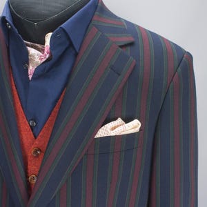 Puede incluir: Un traje a rayas azul marino con rayas burdeos y verdes. El traje incluye una chaqueta a juego, una camisa azul marino, un chaleco color óxido y un pañuelo de bolsillo estampado. El traje se exhibe en una cabeza de maniquí.