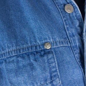 以下が含まれることがあります： 青いデニムジャケットのクローズアップ。ジャケットはシルバー色のボタンとポケットのディテールが特徴です。ステッチが見え、デニムは質感があります。クラシックなスタイルのジャケットです。