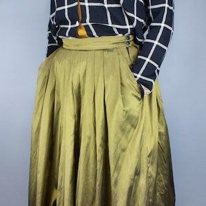 Pode incluir: Uma saia longa verde-oliva com pregas e cintura alta. A saia é feita de um tecido brilhante e tem um efeito degradê sutil.