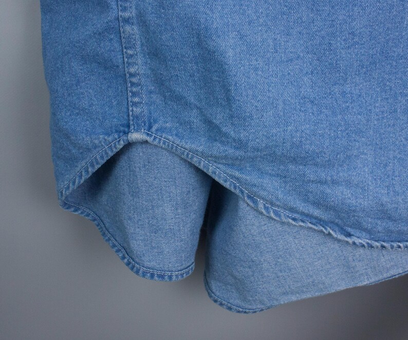 以下が含まれることがあります： 明るいブルーデニムシャツのクローズアップ。画像は、衣服の湾曲した裾とステッチの詳細を強調しています。生地は中程度の重さのデニムのようです。