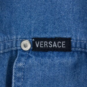 以下が含まれることがあります： 青いデニムの衣服のクローズアップ。黒いラベルには白い文字で「VERSACE」と書かれています。装飾的なデザインのシルバーボタンが見えます。衣服のステッチも確認できます。