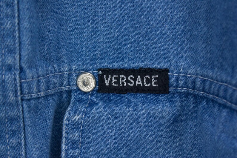 以下が含まれることがあります： 青いデニムの衣服のクローズアップ。黒いラベルには白い文字で「VERSACE」と書かれています。装飾的なデザインのシルバーボタンが見えます。衣服のステッチも確認できます。