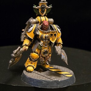 May include: A miniature figurine of a yellow and black armored space marine with a red head and a helmet. The figurine is holding two weapons and stands on a circular base with yellow stripes. The figure has intricate details and is set against a dark background.