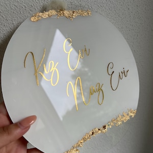May include: A white round acrylic sign with gold lettering that reads "Kız Evi Naz Evi". The sign has gold leaf accents around the edges.