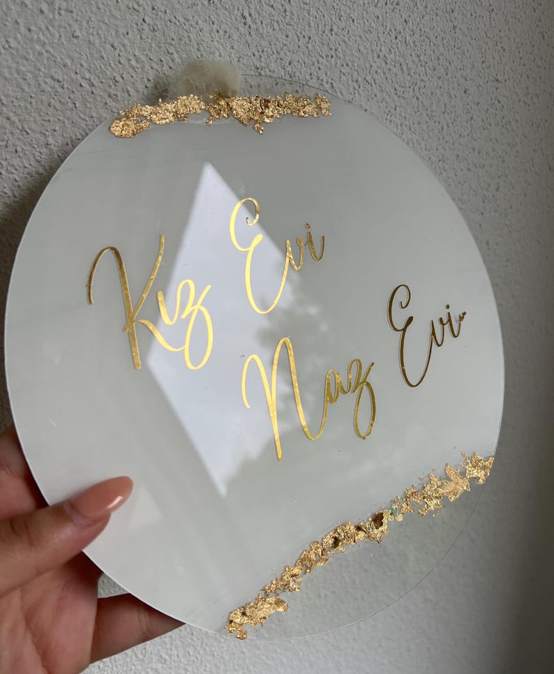 May include: A white round acrylic sign with gold lettering that reads "Kız Evi Naz Evi". The sign has gold leaf accents around the edges.