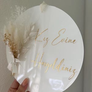 May include: White round sign with gold lettering that says "Kız Evine Hoşgeldiniz". The sign is decorated with a bouquet of dried white and beige pampas grass.