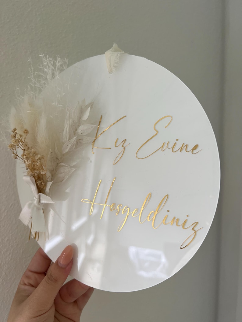 May include: White round sign with gold lettering that says "Kız Evine Hoşgeldiniz". The sign is decorated with a bouquet of dried white and beige pampas grass.