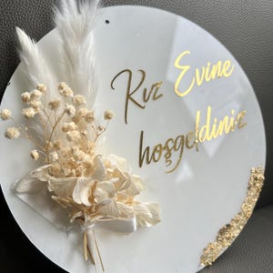 May include: A white round sign with gold lettering that says "Rv Evin hosgeldiniz" and a bouquet of dried white flowers.