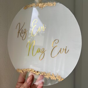 May include: Round white acrylic sign with gold lettering that reads "Kız Evi Naz Evi" and gold leaf accents around the edges.