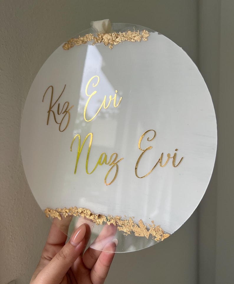May include: Round white acrylic sign with gold lettering that reads "Kız Evi Naz Evi" and gold leaf accents around the edges.